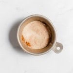 A small metal cup filled with a mixture of finely powdered, light brown and white spices, placed on a white marble surface.