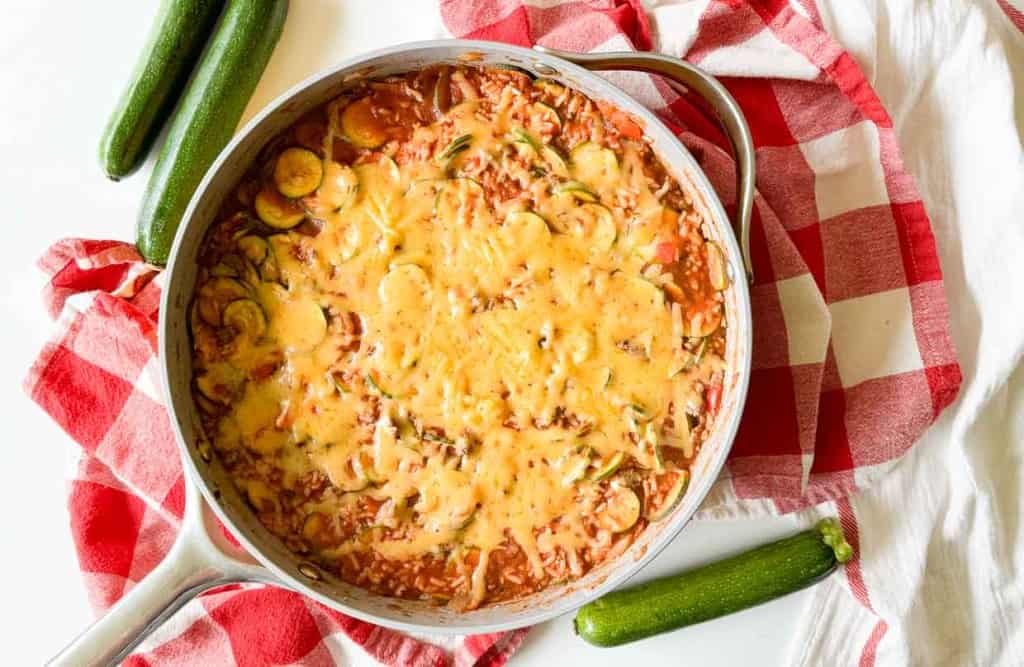 A pan of cheese-topped unstuffed zucchini casserole on a red and white checkered cloth with three whole zucchinis placed around it.