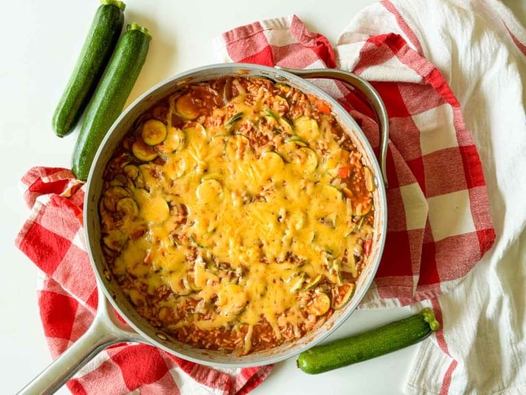 A skillet filled with an unstuffed zucchini casserole topped with melted cheese, placed on a red and white checkered cloth. Whole zucchinis are visible around the skillet.