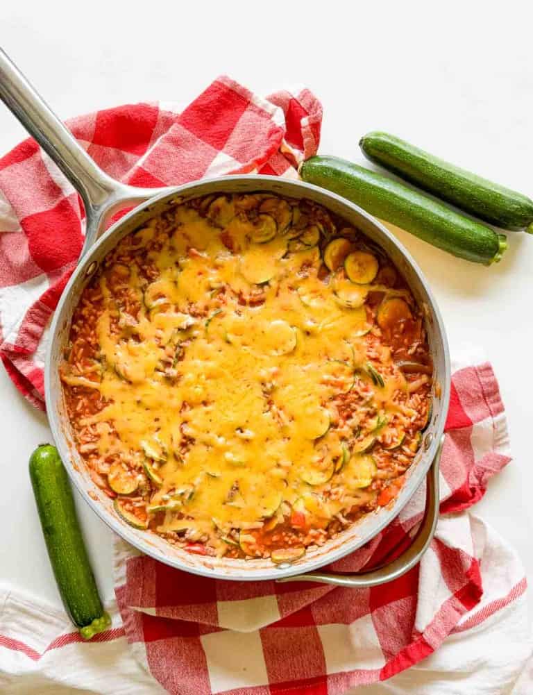 A skillet on top of a red checkered cloth is filled with a delicious unstuffed zucchini casserole, featuring rice and topped with melted cheese. Two whole zucchinis are placed beside the skillet.