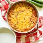 A skillet filled with Unstuffed Zucchini Casserole topped with melted cheese, placed on a red and white checkered cloth next to three zucchinis and an empty white plate.