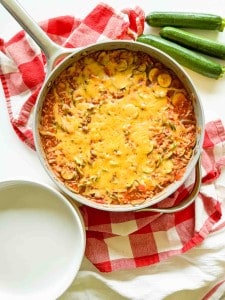 A skillet filled with Unstuffed Zucchini Casserole topped with melted cheese, placed on a red and white checkered cloth next to three zucchinis and an empty white plate.
