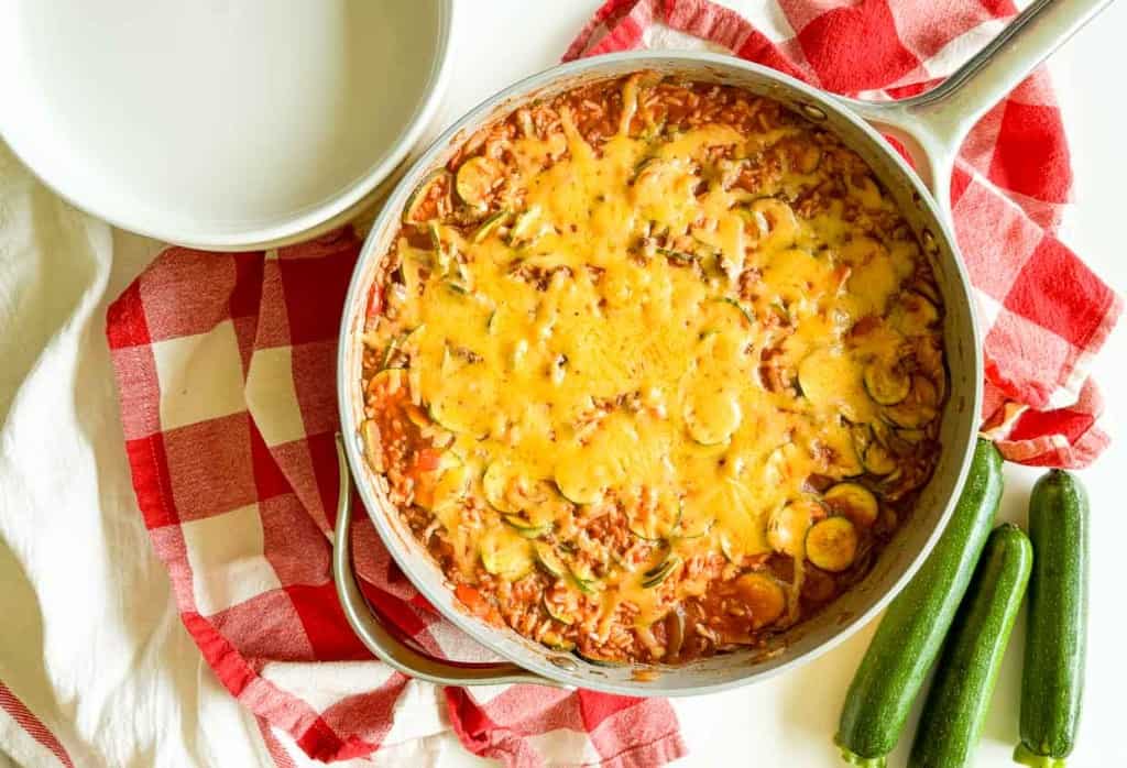 A skillet filled with Unstuffed Zucchini Casserole atop a red and white checkered cloth, with three whole zucchinis beside it. A white bowl is placed near the skillet.