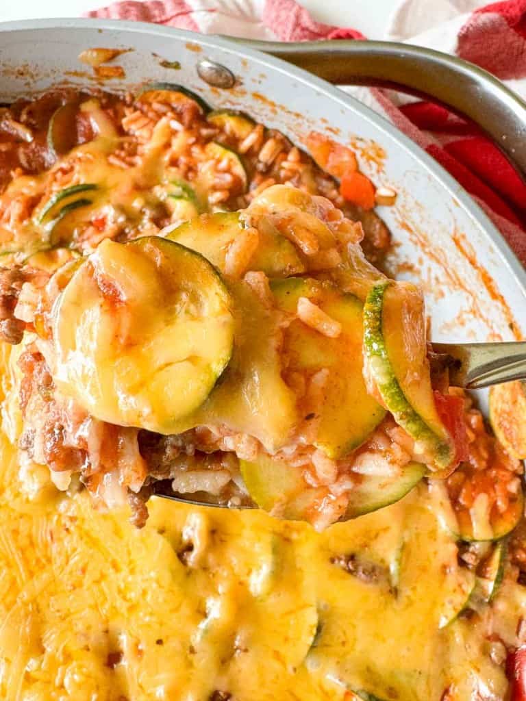 A close-up of an unstuffed zucchini casserole with cheese, rice, and ground meat being scooped from a dish with a metal utensil.