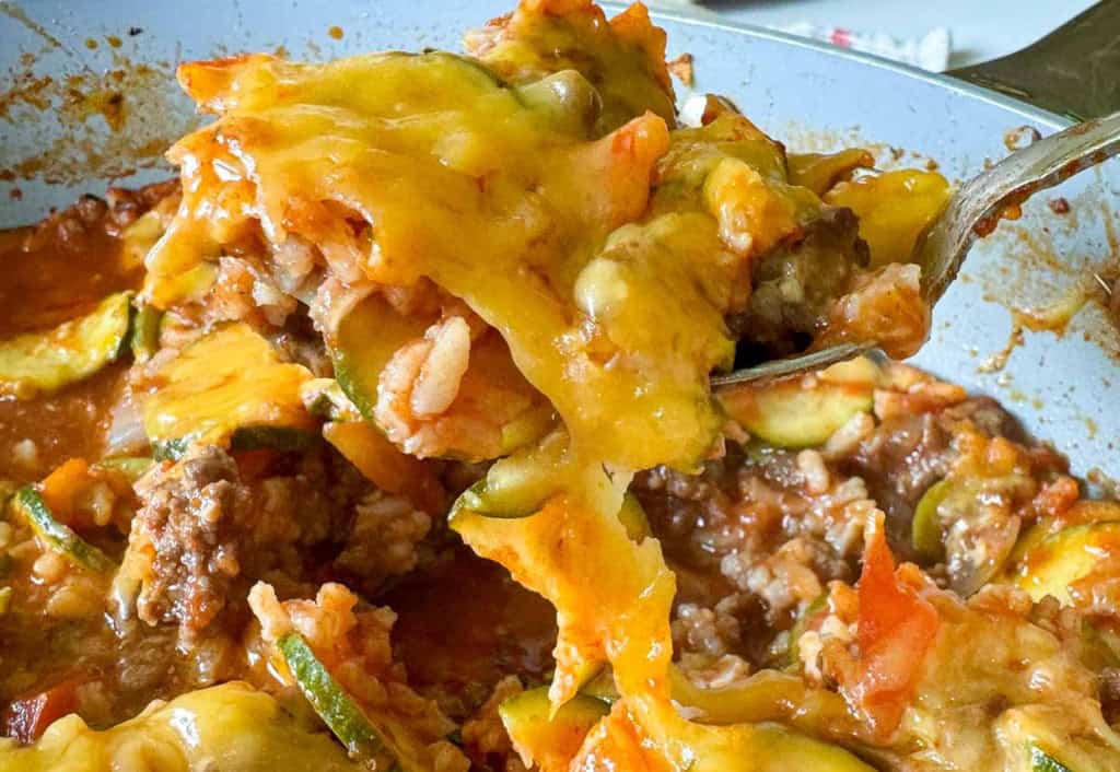 A close-up of a spoon holding a portion of unstuffed zucchini casserole, brimming with cheesy goodness, ground meat, rice, zucchini slices, and rich tomato sauce.