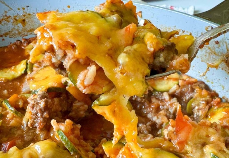 A close-up of a spoon holding a portion of unstuffed zucchini casserole, brimming with cheesy goodness, ground meat, rice, zucchini slices, and rich tomato sauce.