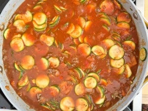 A pan filled with tomato-based sauce, zucchini slices, and ground meat sizzles on the stovetop, creating a delightful Unstuffed Zucchini Casserole.