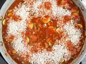 A skillet filled with white rice, tomato sauce, and sliced zucchini being prepared on a stovetop resembles an unstuffed zucchini casserole in the making.
