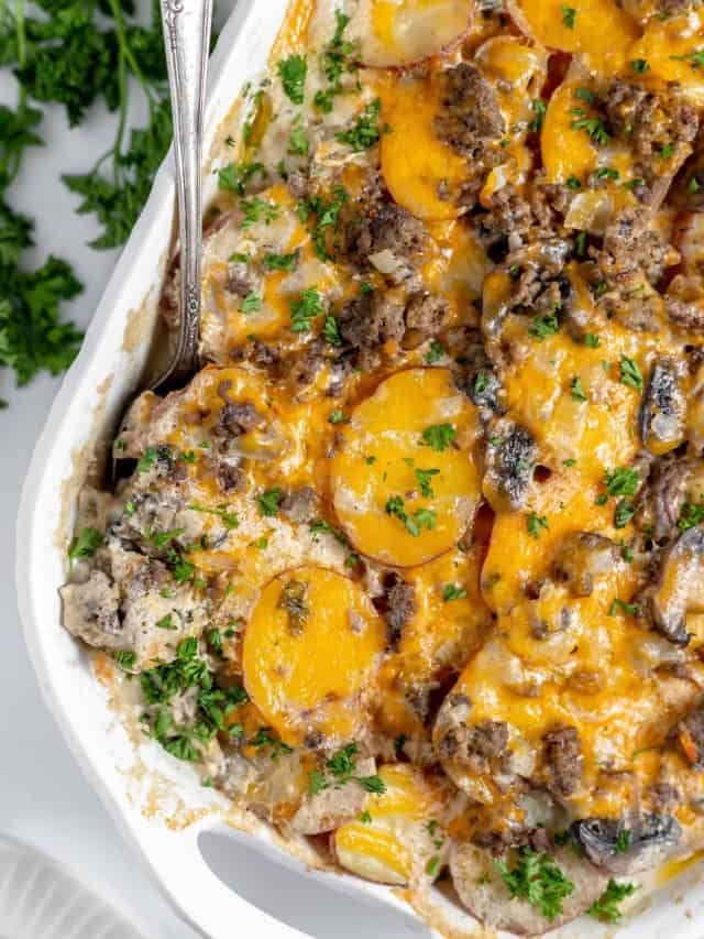 A close-up of a baked casserole with sliced potatoes, ground meat, melted cheese, and sprinkled parsley in a white dish with a spoon.