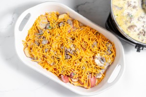 A white baking dish filled with an uncooked mixture of chopped ingredients, topped with shredded cheddar cheese, positioned next to a pot of creamy sauce, makes a perfect base for a delicious Hamburger Potato Casserole.