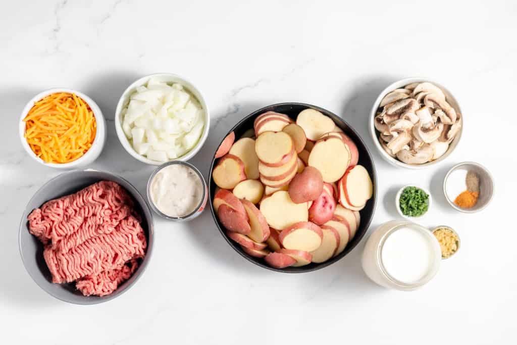 Ingredients arranged on a white surface including ground beef, grated cheese, chopped onions, sliced potatoes, mushrooms, canned soup, chopped herbs, spices, and a cup of milk.