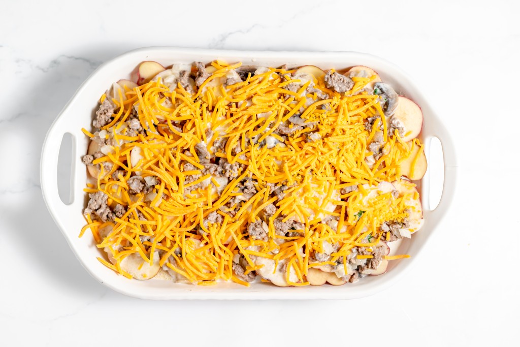 A white oval baking dish filled with a delicious Hamburger Potato Casserole, featuring sliced potatoes, ground meat, and shredded cheese on top, placed on a white countertop.