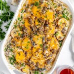 A baked Hamburger Potato Casserole dish with layers of sliced potatoes, cheese, and meat, garnished with chopped parsley. Two glasses of red liquid and some mushrooms are nearby.