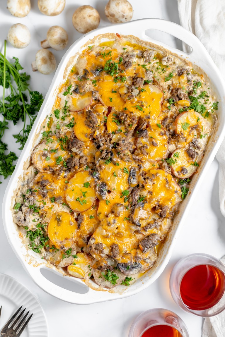 A baked Hamburger Potato Casserole dish with layers of sliced potatoes, cheese, and meat, garnished with chopped parsley. Two glasses of red liquid and some mushrooms are nearby.
