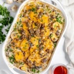 A baked Hamburger Potato Casserole in a white dish, topped with melted cheese, mushrooms, and herbs. A white cloth, parsley, and small bowl of sauce are beside it.