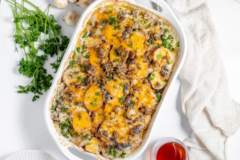 A baked Hamburger Potato Casserole in a white dish, topped with melted cheese, mushrooms, and herbs. A white cloth, parsley, and small bowl of sauce are beside it.