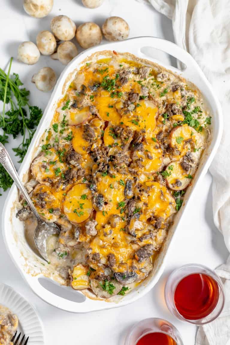 The Hamburger Potato Casserole is a baked dish with cheesy potato slices and ground meat, garnished with chopped parsley. Mushrooms and parsley, along with two glasses of red liquid, are visible nearby.