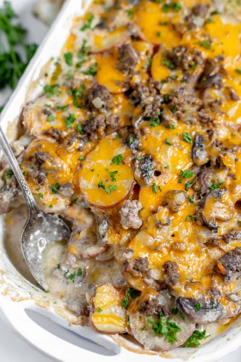 Close-up of a cheesy Hamburger Potato Casserole with ground meat, mushrooms, and chopped parsley on top in a white baking dish, with a serving spoon placed in the dish.