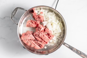 Ground beef and chopped onions cooking in a stainless steel pan on a stovetop.