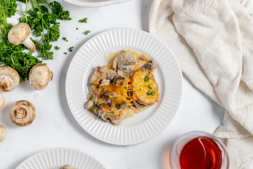 A white plate with a serving of cheesy hamburger potato casserole. Fresh mushrooms, parsley, a white cloth, and a glass of red liquid are around the plate on a white surface.