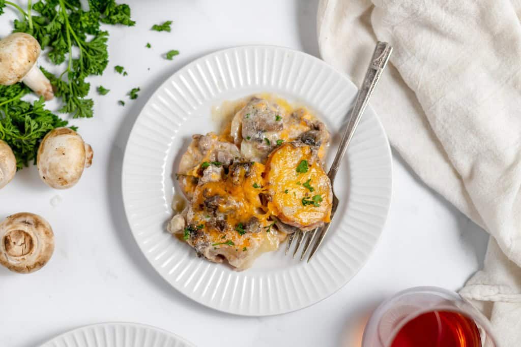 A plate of cheesy scalloped potatoes with mushrooms and herbs, reminiscent of a hearty Hamburger Potato Casserole, accompanied by a fork, a bunch of parsley, whole mushrooms, and a glass of red drink on a white surface.
