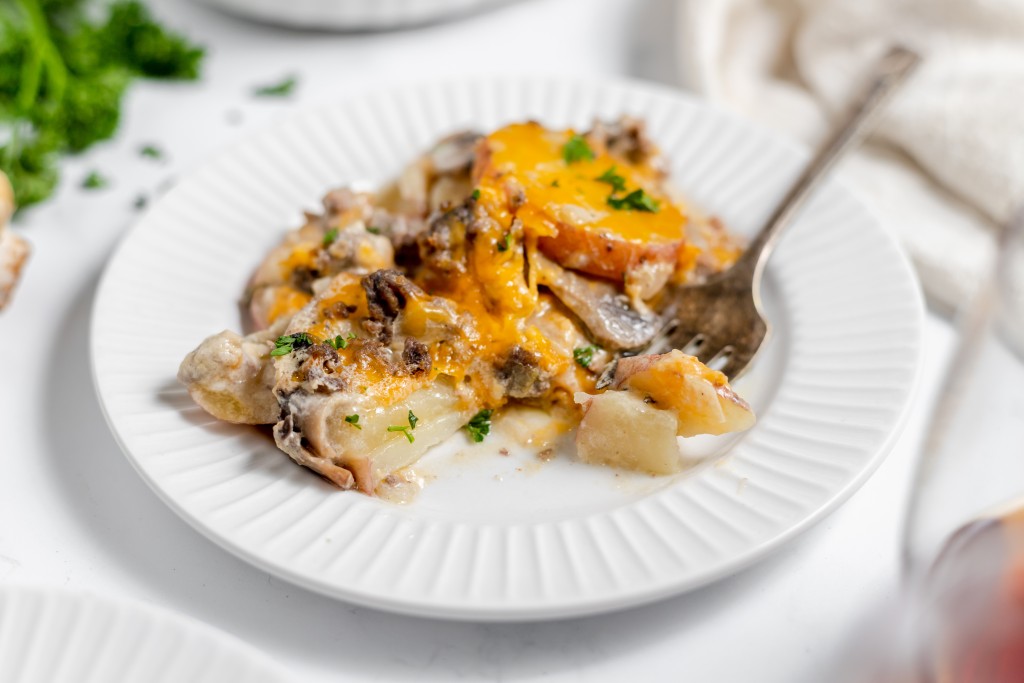 A white plate holds a serving of cheesy potato casserole with a fork on the side, garnished with fresh parsley.