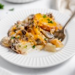 A white plate holds a serving of cheesy potato casserole with a fork on the side, garnished with fresh parsley.