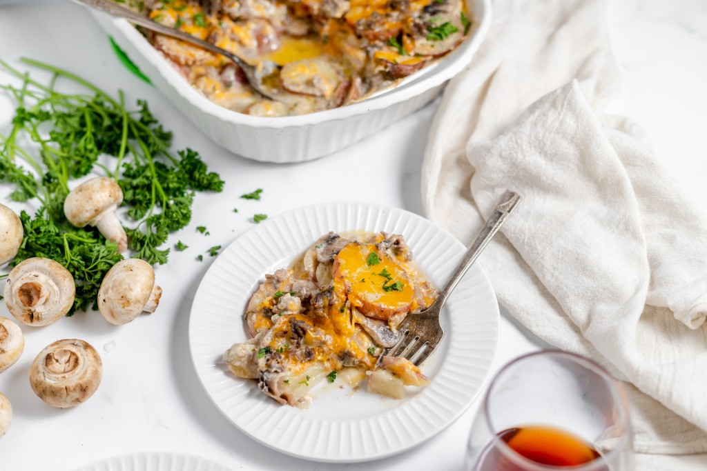 A plate of Hamburger Potato Casserole with mushrooms, cheese, and herbs alongside a glass of drink, mushrooms, and parsley on a white surface.