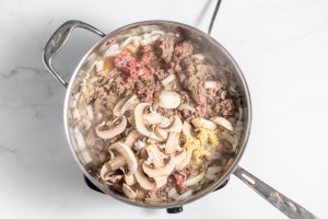 A cooking pot on a stove contains ground meat, sliced onions, minced garlic, and sliced mushrooms being sautéed.