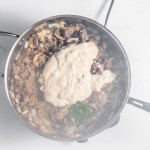 A pot on a stove contains browned ground beef, mushrooms, chopped onions, and a large dollop of creamy white sauce. Steam rises from the mixture, and some herbs are visible.