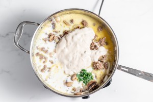 A pot on a stovetop filled with a creamy mixture of ground meat, mushrooms, and herbs, partially covered with a dollop of cream or sauce.