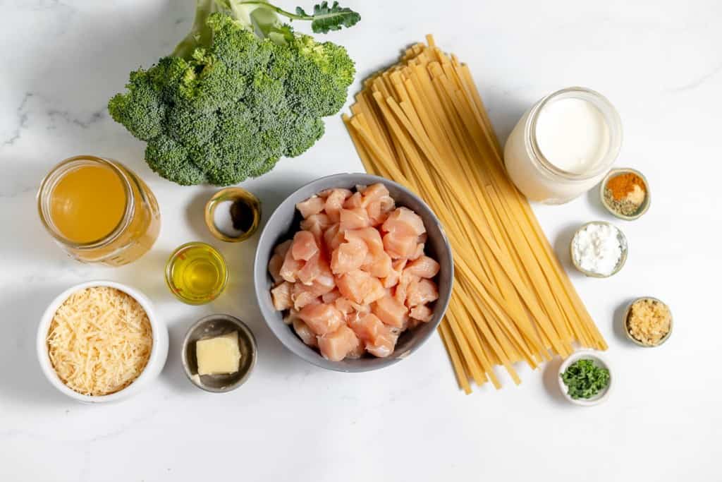 Ingredients for a meal displayed on a countertop, including broccoli, uncooked pasta, diced raw chicken, cream, grated cheese, olive oil, butter, garlic, cornstarch, broth, and seasonings in small bowls.