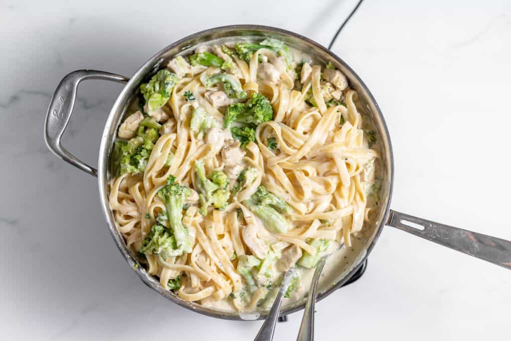 A stainless steel pan filled with creamy fettuccine Alfredo pasta mixed with broccoli and chicken pieces on a white marble surface.