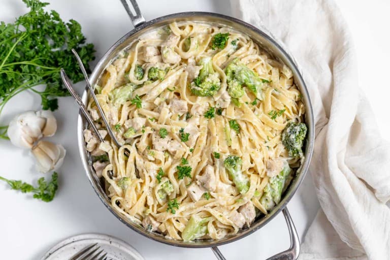 A pan filled with creamy chicken and broccoli fettuccine Alfredo, garnished with parsley. A serving spoon is placed in the pan, and fresh parsley and garlic bulbs are seen beside it.