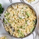 A stainless steel pan filled with fettuccine Alfredo pasta, broccoli florets, and chicken pieces, garnished with chopped parsley. Two servings of the dish are plated on the side.