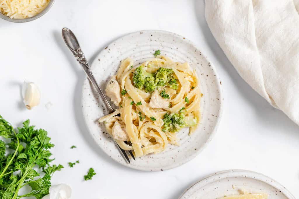 A plate of fettuccine pasta with chicken and broccoli, garnished with parsley. A fork rests on the plate. Fresh parsley, a garlic clove, and a white cloth are nearby.