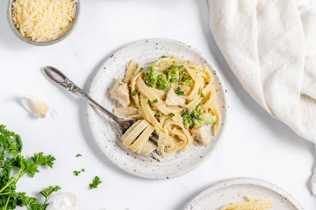 A white plate with fettuccine pasta, broccoli, and a creamy sauce, garnished with parsley. A fork is placed on the plate. Surrounding the plate are a cloth napkin, a bowl of grated cheese, parsley, and garlic cloves.