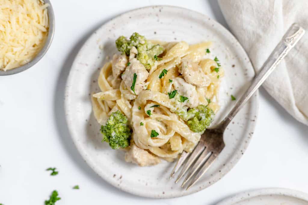 A plate of fettuccine pasta with creamy white sauce, chicken pieces, and broccoli, garnished with parsley. A fork is placed beside the dish and a bowl of grated cheese is partially visible.