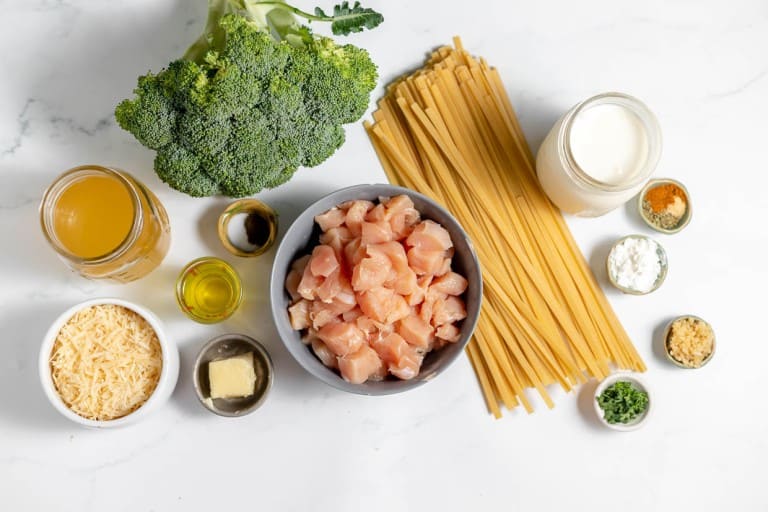 Ingredients for a meal displayed on a countertop, including broccoli, uncooked pasta, diced raw chicken, cream, grated cheese, olive oil, butter, garlic, cornstarch, broth, and seasonings in small bowls.