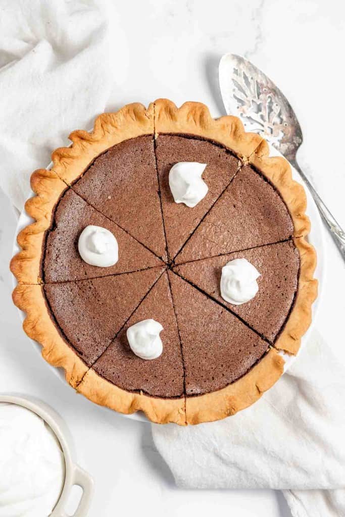 A sliced chocolate pie with dollops of whipped cream on top, placed on a white cloth, next to a bowl of whipped cream and a pie server.
