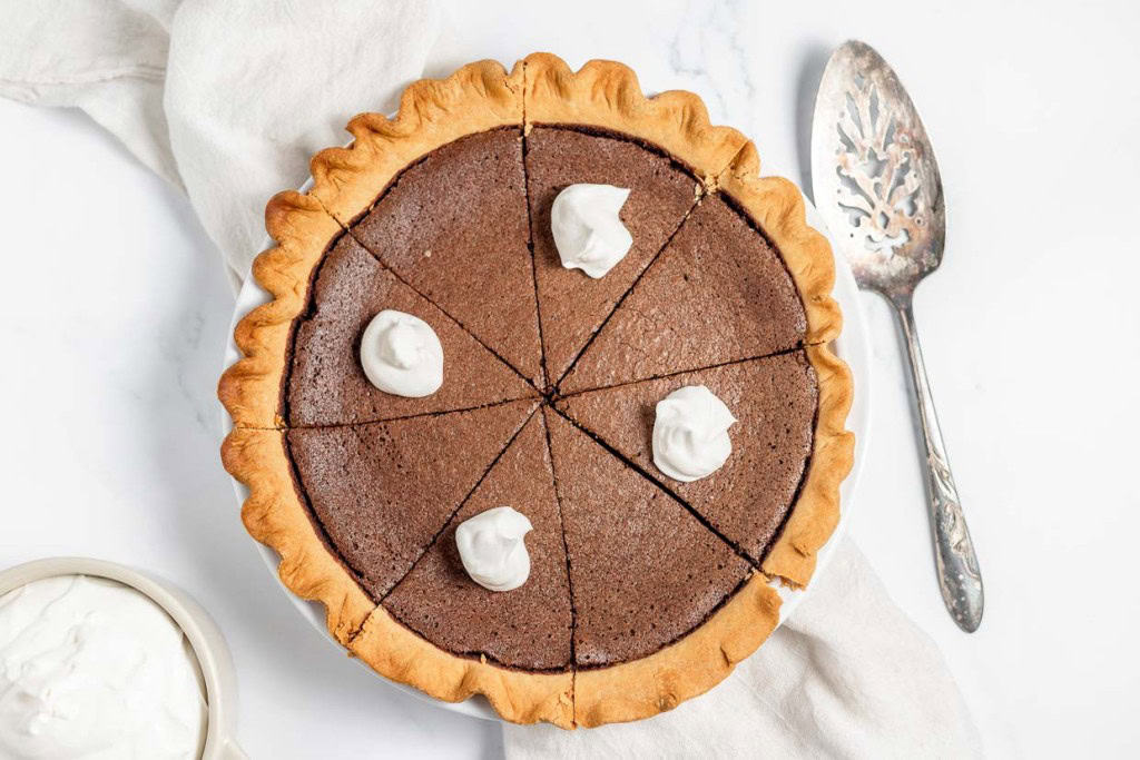A chocolate pie cut into six slices with dollops of whipped cream on top, placed on a white cloth next to a silver pie server and a bowl of whipped cream.