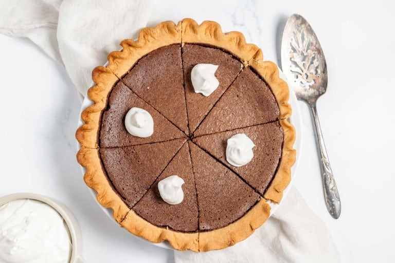 A chocolate pie cut into six slices with dollops of whipped cream on top, placed on a white cloth next to a silver pie server and a bowl of whipped cream.