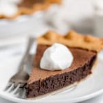 A slice of chocolate pie topped with a dollop of whipped cream is placed on a white plate with a fork beside it.