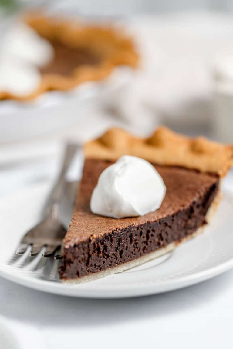 A slice of chocolate pie topped with a dollop of whipped cream is placed on a white plate with a fork beside it.
