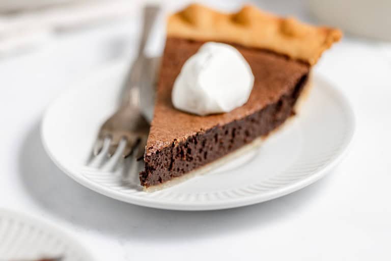 A slice of chocolate pie topped with a dollop of whipped cream on a white plate with a fork and knife.