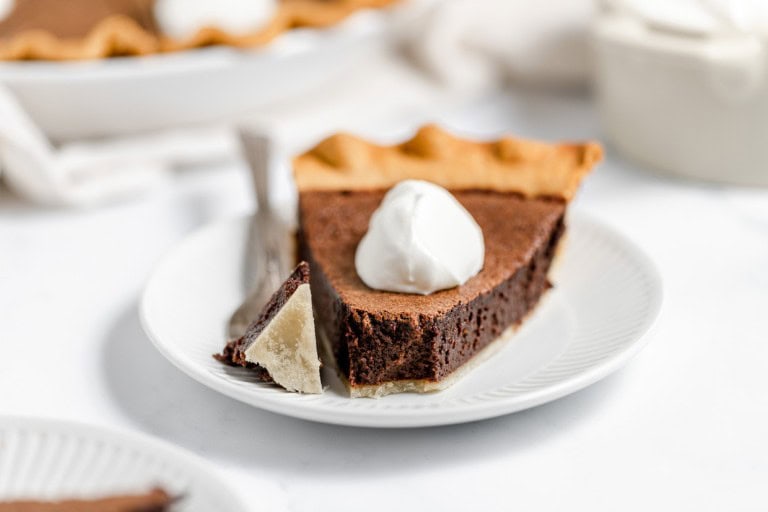 A slice of chocolate pie topped with whipped cream on a white plate, with a fork holding a piece of the pie.