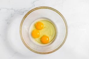 A clear glass bowl containing three raw egg yolks and some egg whites, placed on a white marble surface.