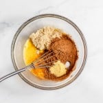 A glass mixing bowl containing unmixed ingredients: brown sugar, cocoa powder, melted butter, and vanilla extract with a metal whisk on a white marble surface.