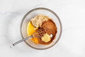 A glass mixing bowl containing unmixed ingredients: brown sugar, cocoa powder, melted butter, and vanilla extract with a metal whisk on a white marble surface.