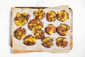 A baking tray lined with parchment paper holds several smashed red potatoes seasoned with herbs and roasted to a golden finish.